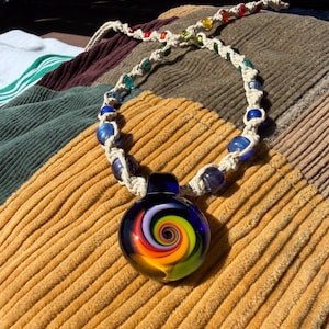 May include: A handmade necklace featuring a dark blue glass pendant with a colorful spiral design. The pendant is attached to a cream-colored rope necklace with blue and colorful glass beads. The necklace rests on a corduroy surface.