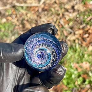 Puede incluir: Una canica de vidrio azul con un diseño de galaxia en espiral y detalles de purpurina plateada.