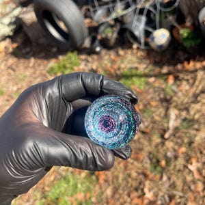 Puede incluir: Un pequeño orbe de vidrio redondo con un diseño de espiral iridiscente en tonos azules, morados y verdes. El orbe está cubierto de purpurina.