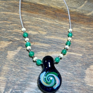 May include: A handcrafted necklace featuring a dark blue glass pendant with a colorful spiral design. The pendant is suspended from a silver chain, accented with green and off-white beads. The necklace is displayed on a wooden surface.