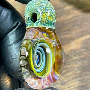 May include: A glass pendant with a swirling yellow, blue, and white pattern inside. The top of the pendant is green and brown with a small hole in the center.