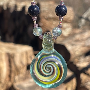 May include: A glass pendant necklace with a swirling, multi-colored design. The pendant is a teardrop shape and is suspended from a beaded chain with turquoise and black beads.