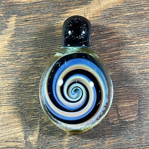May include: A glass pendant with a black, white, and brown spiral design. The pendant has a black top and a clear bottom.