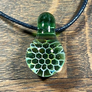 Puede incluir: Un colgante de vidrio verde con un patrón de panal. El colgante está en un cordón negro.