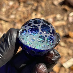 May include: A clear glass sphere with a pattern of various sized circles. The sphere is filled with blue and silver glitter. The sphere is held by a person wearing black gloves. The background is blurred.