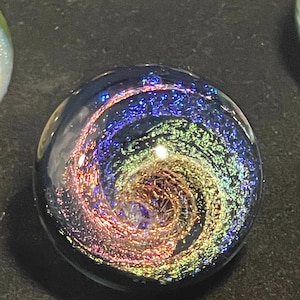 Puede incluir: Una canica de vidrio con un diseño de galaxia en espiral en tonos de azul, verde, rosa y dorado. La canica tiene un fondo negro.