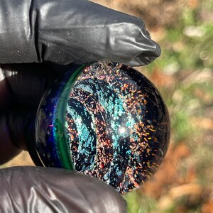May include: A dark blue glass marble with iridescent green, blue, and orange flecks. The marble has a swirling pattern and a high gloss finish.