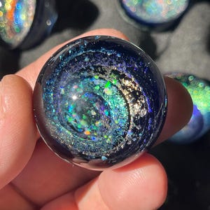 Puede incluir: Un globo de cristal transparente con un diseño de galaxia arremolinada. El globo presenta tonos de azul, verde y purpurina plateada, creando un efecto cósmico. Otros globos similares son visibles en el fondo.