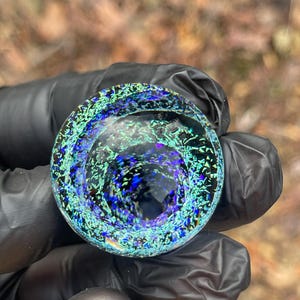 Puede incluir: Un pisapapeles redondo de vidrio azul y verde con un patrón de remolino, como una galaxia. El pisapapeles es translúcido y tiene un centro azul oscuro.
