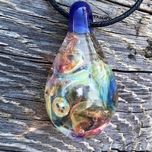 Peut inclure: Un pendentif en verre en forme de larme avec un dessus bleu et des motifs tourbillonnants de blanc, jaune, orange et rose à l'intérieur. Le pendentif est suspendu à un cordon noir.
