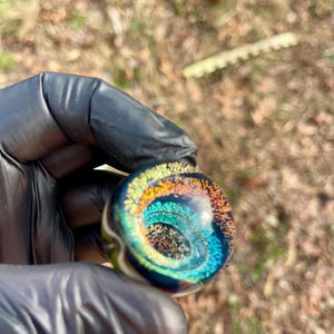 Puede incluir: Una canica de vidrio redonda con un patrón de espiral de azul, verde, naranja y amarillo. La canica está llena de pequeñas partículas iridiscentes.