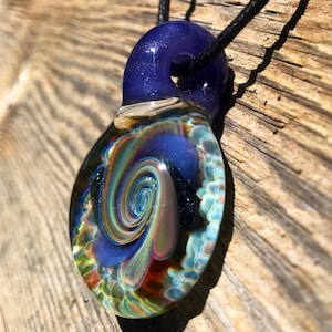 May include: A glass pendant necklace with a swirling blue, green, and gold design. The pendant is attached to a black cord.
