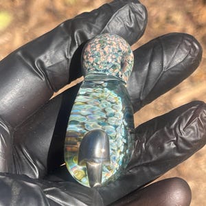 Könnte beinhalten: Ein Glasanhänger mit einem Pilzdesign. Die Oberseite des Anhängers ist mit rosa und blaugrünen Flecken versehen. Der Körper ist transparent mit farbenfrohen, schillernden Akzenten. In der Mitte befindet sich eine silberne Pilzform.