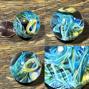 Puede incluir: Una canica de vidrio azul y amarillo con patrones de remolino y un centro transparente. La canica tiene un anillo amarillo en el interior.