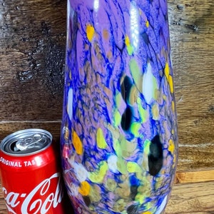 May include: A tall, cylindrical glass jar with a cork lid. The jar is decorated with a swirling pattern of purple, blue, yellow, green, and white. The jar is sitting on a wooden surface.