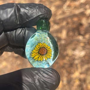 Puede incluir: Colgante de cristal transparente con un diseño de girasol amarillo. El colgante tiene un fondo azul claro y blanco, con una parte superior verde. El girasol tiene un centro marrón y pétalos amarillos.