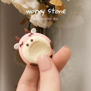 Könnte beinhalten: Ein rosa Sorgenstein aus Polymer Clay in Kuhform. Er hat braune Flecken und Hörner sowie eine cremefarbene Mitte. Der Text auf dem Bild lautet "worry stone out of polymer clay."