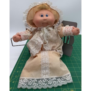 May include: A vintage doll with a round face, blue eyes, and a peach-colored complexion. The doll wears a peach-colored dress and bonnet, both trimmed with white lace. The doll is seated in a small, gray chair.