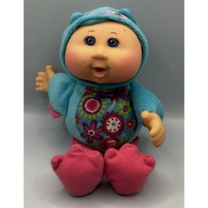 May include: A Cabbage Patch Kids doll wearing a blue hooded outfit with a floral design on the front and pink feet. The doll has blue eyes and rosy cheeks. The doll is sitting and has its right arm raised.