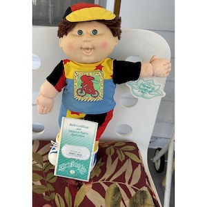 Peut inclure: Poupée Cabbage Patch Kids vêtue d'une tenue jaune, bleue, noire et rouge avec une casquette assortie. La poupée a les yeux verts et des taches de rousseur. Elle tient un certificat de naissance et des papiers d'adoption. Une étiquette indique "J'adore rire aux blagues."