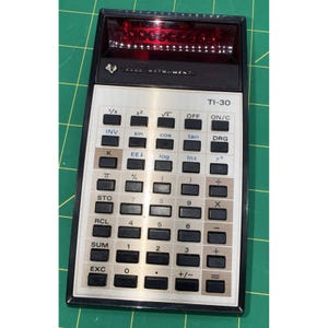 Könnte beinhalten: Ein Vintage Texas Instruments TI-30 Taschenrechner. Der Rechner hat ein schwarzes und silbernes Gehäuse mit einer roten LED-Anzeige. Er verfügt über zahlreiche Tasten für wissenschaftliche Berechnungen, einschließlich trigonometrischer Funktionen, Logarithmen und Exponenten. Der Rechner ist für den Bildungs- und professionellen Gebrauch konzipiert.
