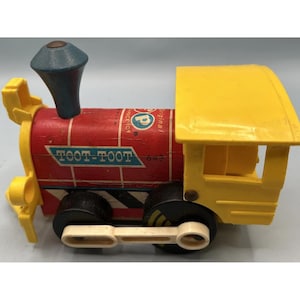 May include: A vintage Fisher-Price toy train engine featuring a red cylindrical body with white grid lines, a blue smokestack, and a yellow cab. The side displays the text TOOT-TOOT 643 and the Fisher-Price logo. It has black wheels and white plastic connecting rods.