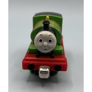 May include: A toy train with a smiling face, green body, and red base. The train has black wheels and a silver-coloured front bumper. The top of the train is black, and the face is white with black eyes.