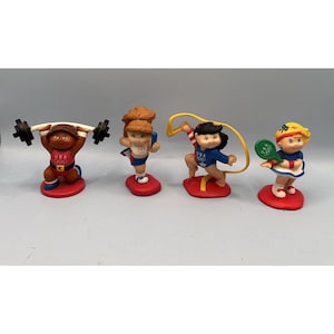 May include: A collection of four Olympic-themed figurines. The figurines depict athletes in various sports, including weightlifting, gymnastics, and tennis. Each figurine is on a red base and wears a USA uniform.