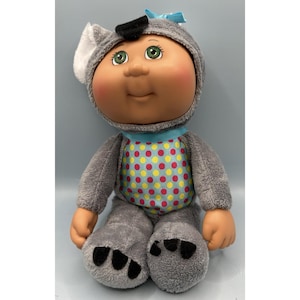 May include: A Cabbage Patch Kids doll wearing a gray koala-themed outfit. The doll has green eyes, rosy cheeks, and a blue bow. The outfit includes a blue and polka-dotted shirt and koala feet.