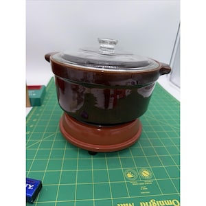May include: A brown ceramic casserole dish with a clear glass lid. The dish sits on a matching brown warming base. The casserole dish has two handles and is designed for cooking and serving food.
