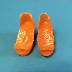 May include: A pair of orange doll shoes with open toes. The shoes are made of a solid material and have a simple, classic design. The shoes are set against a blue background.