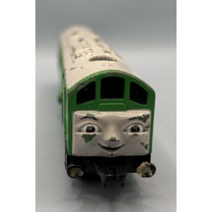 May include: A vintage toy train with a smiling face, painted in white and green. The toy train has a detailed design with visible wear and tear, suggesting it's a well-loved collectible. The train's face has a friendly expression.