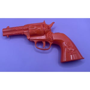 Könnte beinhalten: Ein orangefarbener Spielzeugrevolver aus Kunststoff mit detaillierten, geprägten Ornamenten an Lauf und Trommel, einem Relief eines Reiters auf dem Griffstück und dem deutlich sichtbaren Schriftzug "Dragon-1" über dem Abzugsbügel.