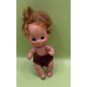 May include: A vintage doll with a peach skin tone, blue eyes, and reddish-brown hair. The doll is wearing brown shorts. The doll has rosy cheeks and is posed in a standing position.
