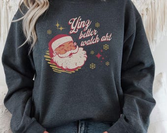 May include: Dark gray sweatshirt featuring a Santa Claus graphic with the text "Yinz better watch aht" in a retro font. The design includes snowflakes and stars, creating a festive holiday theme.