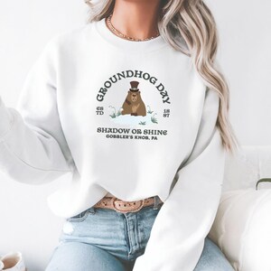 May include: White sweatshirt featuring a Groundhog Day graphic. The design includes a groundhog wearing a top hat, the text "GROUNDHOG DAY", "SHADOW OR SHINE", and "GOBBLER'S KNOB, PA". The sweatshirt has long sleeves and a crew neck.