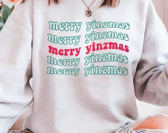 May include: A light gray sweatshirt with the text "merry yinzmas" in green and red. The text is stacked in a repeating pattern. The sweatshirt is a casual style.