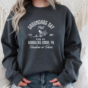 May include: Dark gray sweatshirt with white text and a Groundhog Day graphic. The text includes "GROUNDHOG DAY Phil", "EST. FEB 02 1887", and "GOBBLERS KNOB, PA Shadow or Shine". The design features a groundhog and a top hat.