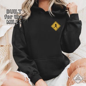 May include: Black hoodie with the text "BUILT for the MILES" and a yellow diamond-shaped patch with a running figure. The hoodie has a front pocket and a hood. The image also shows a person wearing the hoodie.