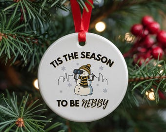 May include: White ceramic Christmas ornament with a red ribbon, featuring a snowman with binoculars, a yellow and black striped scarf and hat, and the text "Tis the Season to be Nebby". Snowflakes and a city skyline are also included.