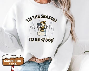 May include: White sweatshirt with a graphic of a snowman wearing a hat and scarf, holding binoculars. The text reads "Tis the Season to be Nebby" with snowflakes and a city skyline. The sweatshirt is a winter-themed apparel item.