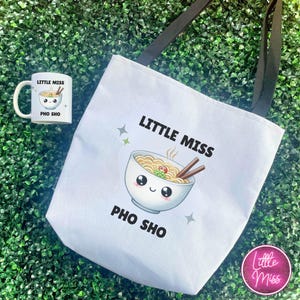 May include: White tote bag and mug featuring a cartoon bowl of pho with chopsticks. The text "LITTLE MISS PHO SHO" is printed on the tote bag and mug. The tote bag has black straps and a pink "Little Miss" logo.