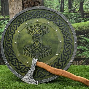 Könnte beinhalten: Ein grünes und schwarzes Wikingerschild mit einem Thorhammer-Design und einem keltischen Knotenmuster. Eine Axt mit Holzgriff liegt auf dem Gras. Der Schild hat einen Durchmesser von etwa 76 cm.