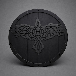 Pode incluir: Um escudo redondo de madeira preta com um design detalhado de corvo no centro. O corvo &eacute; intrincadamente esculpido com um padr&atilde;o de n&oacute; celta. O escudo tem uma borda de metal e &eacute; cravejado com pequenos rebites.