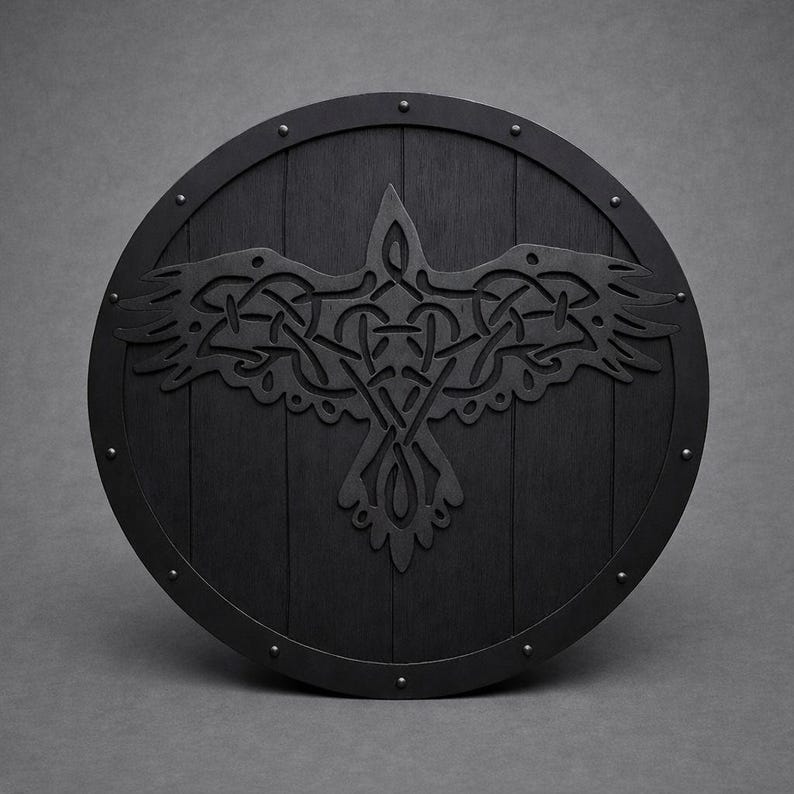 Pode incluir: Um escudo redondo de madeira preta com um design detalhado de corvo no centro. O corvo &eacute; intrincadamente esculpido com um padr&atilde;o de n&oacute; celta. O escudo tem uma borda de metal e &eacute; cravejado com pequenos rebites.