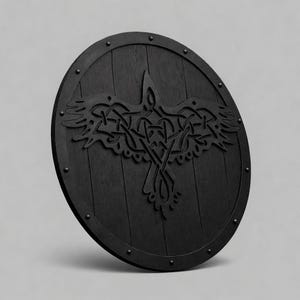 Pode incluir: Um escudo de madeira redondo e preto com um desenho de corvo esculpido. O escudo apresenta um acabamento escuro, tachas de metal ao redor da borda e um motivo de corvo detalhado no centro. O desenho do corvo &eacute; um padr&atilde;o de n&oacute;s complexo.