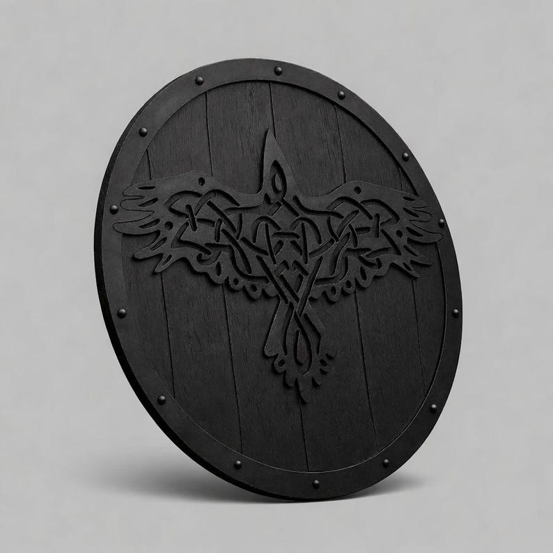 Pode incluir: Um escudo de madeira redondo e preto com um desenho de corvo esculpido. O escudo apresenta um acabamento escuro, tachas de metal ao redor da borda e um motivo de corvo detalhado no centro. O desenho do corvo &eacute; um padr&atilde;o de n&oacute;s complexo.