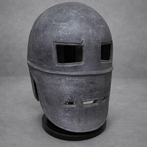 以下が含まれることがあります： 長方形の目の開口部と口のスリットを備えた、グレーの金属製ヘルメット。ヘルメットはテクスチャ加工された表面を持ち、黒いベースに取り付けられています。デザインは、スーパーヒーローやSFの美学を彷彿とさせます。