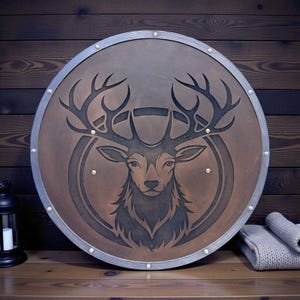 Könnte beinhalten: Ein runder, brauner Schild mit silberner Umrandung und Nieten. Der Schild zeigt eine detaillierte Gravur eines Hirschkopfes mit großem Geweih. Der Hirsch ist in einem kreisförmigen Design zentriert. Der Schild wird auf einer Holzoberfläche präsentiert.