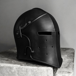 May include: A matte black metal knight helmet featuring a horizontal eye slit and a cross-shaped ventilation cutout on the front face. The helmet includes decorative metal hardware, rivets, and a small chain detail attached to the side panel.